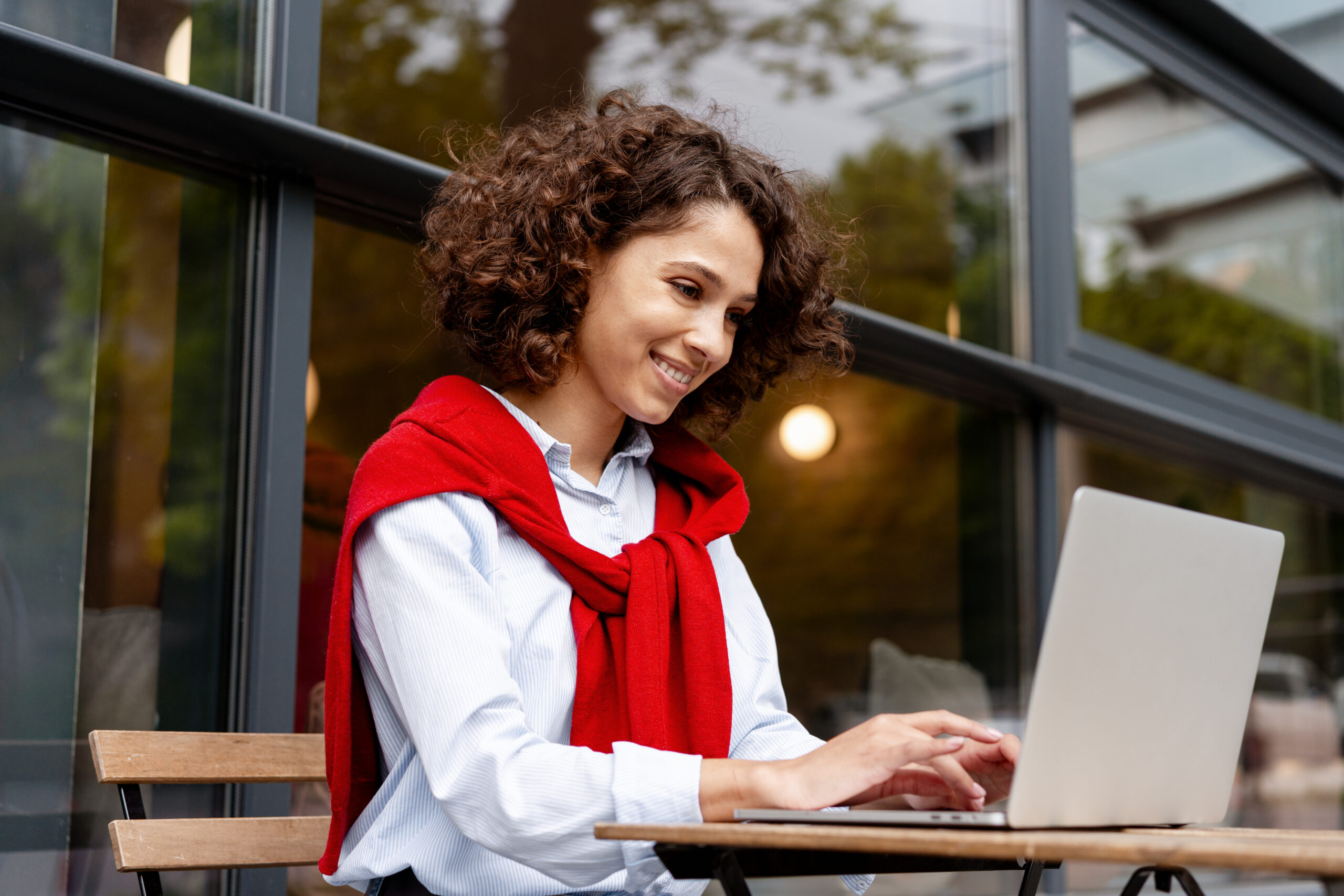 smiling woman working on laptop at outdoor cafe wearing casual red sweater